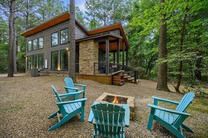 Une cabane moderne avec des fondations en pierre, de grandes fenêtres et une terrasse. On peut voir au premier plan des fauteuils Adirondack et un foyer.