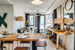 A modern dining area with a wooden table set for a meal, a sofa, and a large window offering a view of the city.