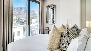 A hotel room with a large bed, a view of a cityscape, a mirror, and a chair. - Beautiful Apartment with Mountain Views (404 on N) (Cape Town)
