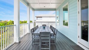 A covered outdoor deck with a table and chairs, a white railing, and a view of a house in the background. - Private Pool | Golf Carts | EV Charger | Elevator | Pet-Friendly (Gulf Shores)