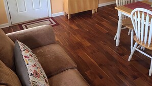 A living room with a sofa, a wooden TV stand, a dining table with chairs, a window with blinds, and a door with curtains. - One Bedroom One Bath Apartment (Selmer)