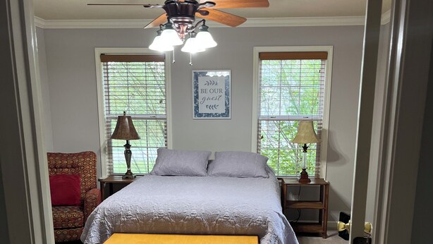 A bedroom with a bed, a ceiling fan, two bedside lamps, a bench, and a wall-mounted air conditioning unit. - One Bedroom One Bath Apartment (Selmer)