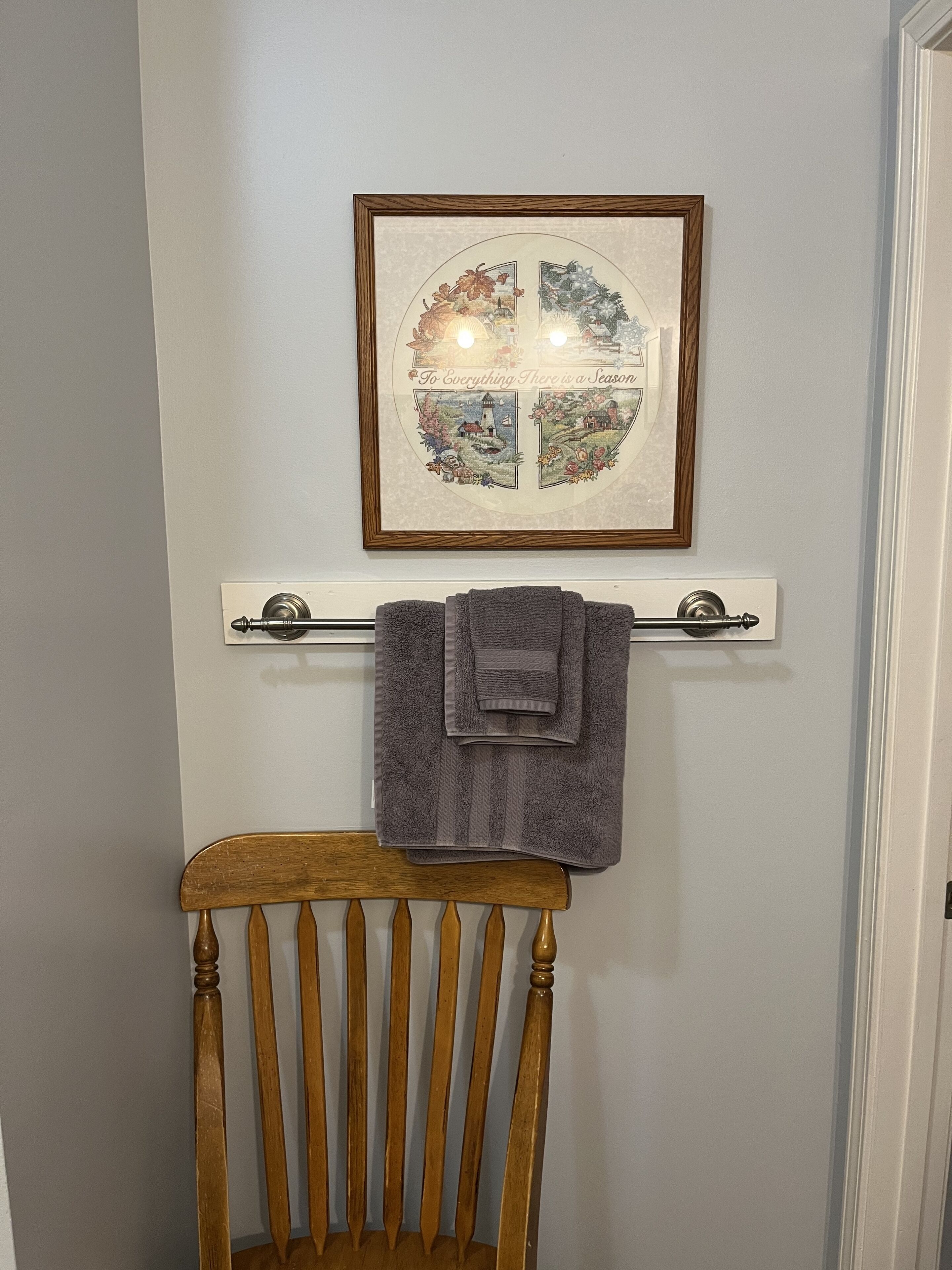 Una obra de arte enmarco que muestra escenas de las estaciones sobre un toallero con una toalla doblada, una silla de madera debajo y una puerta blanca a la derecha.