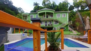 Un área de piscina con cubierta de madera, un edificio verde al fondo y una escalera que lleva a un balcón.