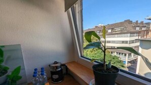 Un pequeño balcón con una planta en maceta, una ventana con vistas a edificios, y una mesa con agua embotellada y una cafetera.