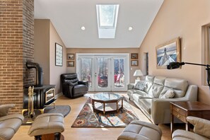 A living room with a fireplace, leather furniture, a glass coffee table, and a view through glass doors. - Spacious Trickey Moose Family Retreat - Woodstove, Firepit, Kayaks, Water Access (Naples)