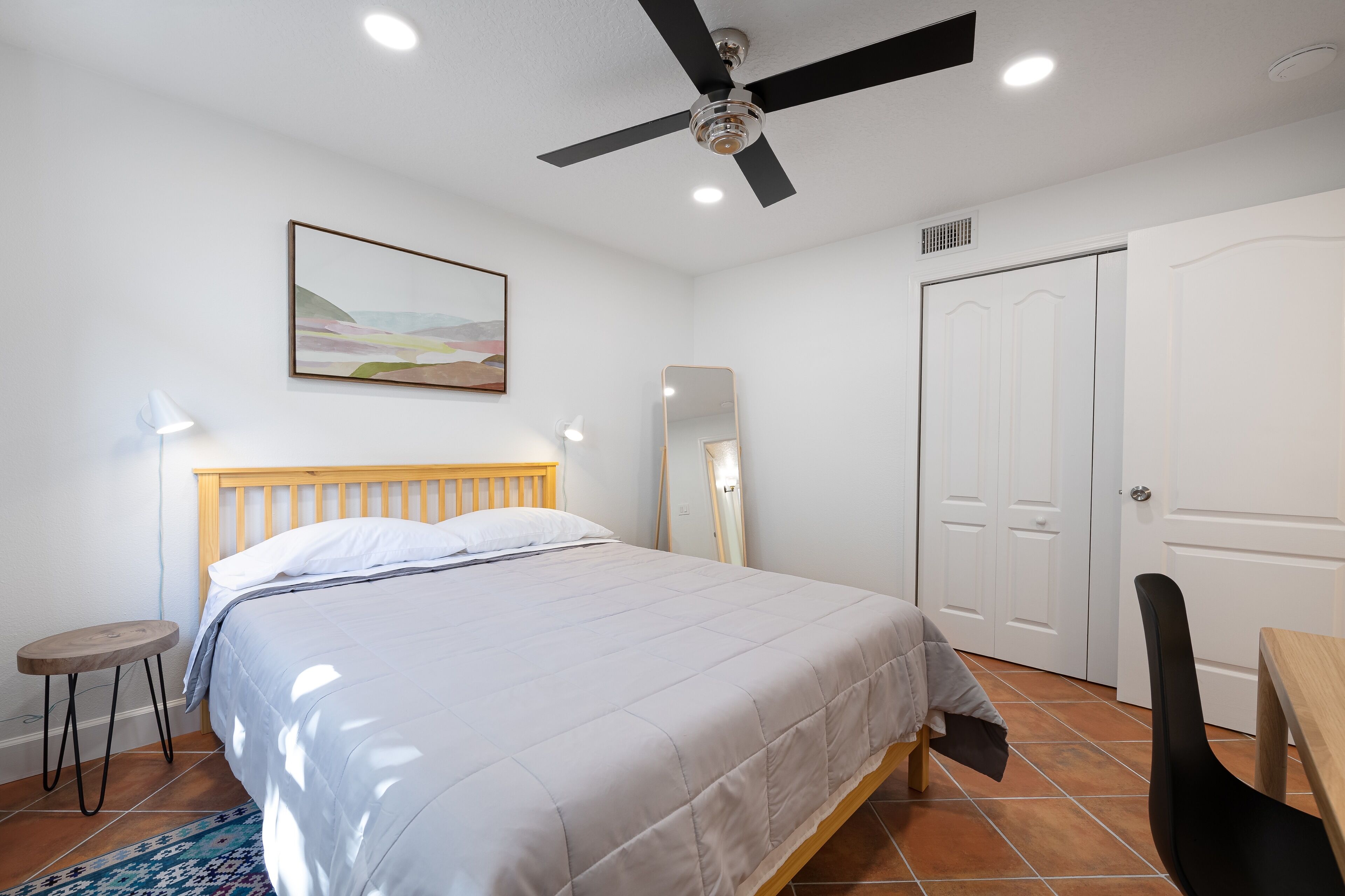 A bedroom with a bed, a ceiling fan, a bedside table, a chair, a desk, a mirror, and a door.