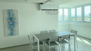 A modern dining area with a white table and chairs, a ceiling-mounted air conditioner, and a large window with a city view.