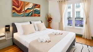 A neatly made bed with rolled towels, a wall art piece, a vase with dried flowers, and a window with curtains.