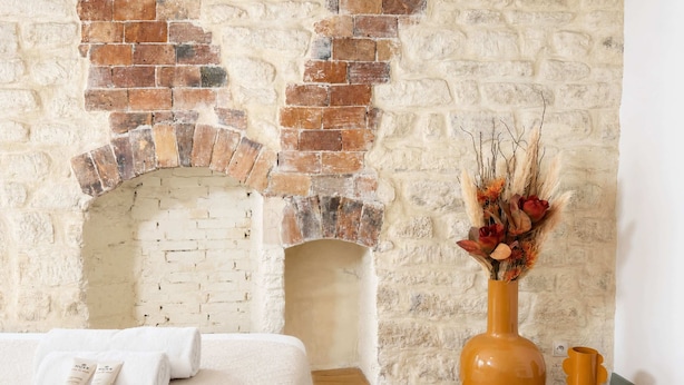A room with a brick fireplace, a bed with white bedding, and a vase with dried flowers on a wooden table. - Authentic parisian property in the Marais (Paris)