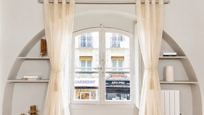 A room with a large arched window, white curtains, and wooden shelves with books and decorative items. - Authentic parisian property in the Marais (Paris)