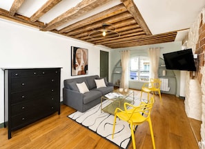 A living room with exposed wooden beams, a sofa, a glass coffee table, a yellow chair, a black dresser, and a TV mounted on the wall. - Authentic parisian property in the Marais (Paris)