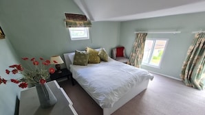 Une chambre à coucher avec un lit, une table de chevet, une lampe, un vase avec des fleurs et une fenêtre avec des rideaux.