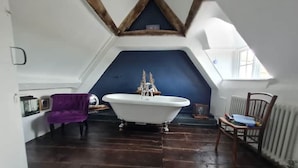 Une pièce avec un plafond en pente, une baignoire blanche, un plancher en bois et une chaise violette.