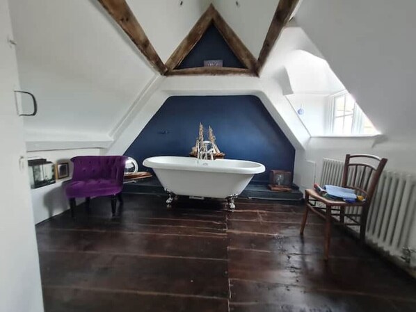 A room with a sloped ceiling, a white bathtub, wooden flooring, and a purple chair.