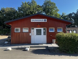 Ein Saunagebäude mit roter Fassade, einer weißen Tür und einem Schild mit der Aufschrift 'SAUNAHAUS'.