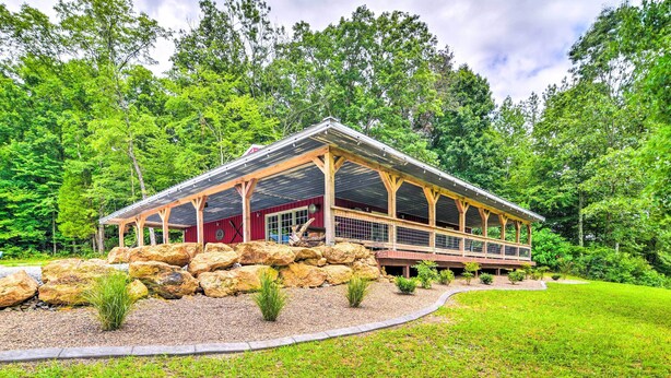 Uma cabana com deck coberto e fundação de pedra, rodeada por um gramado bem cuidado e uma floresta densa.