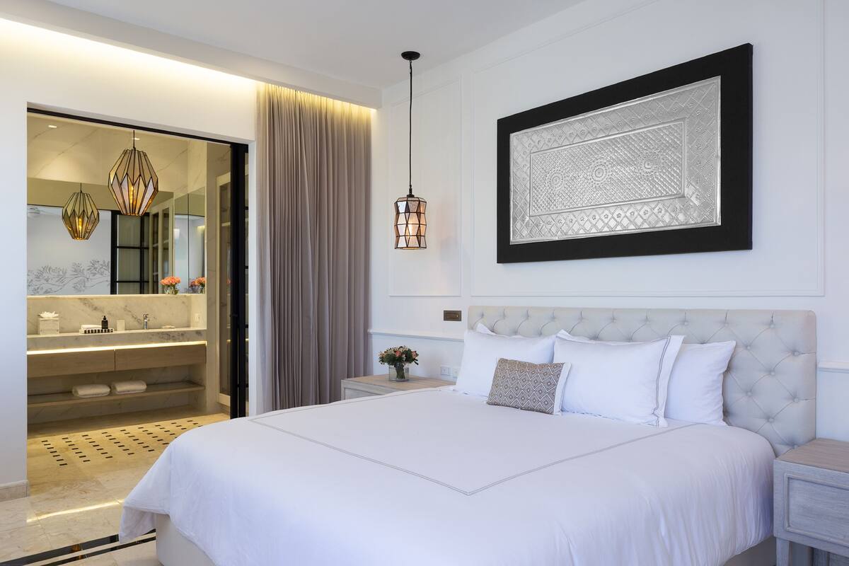 Un dormitorio moderno con una cama grande, una mesita de noche, una lámpara de noche, una obra de arte enmarcada y un baño visible a través de una puerta abierta.
