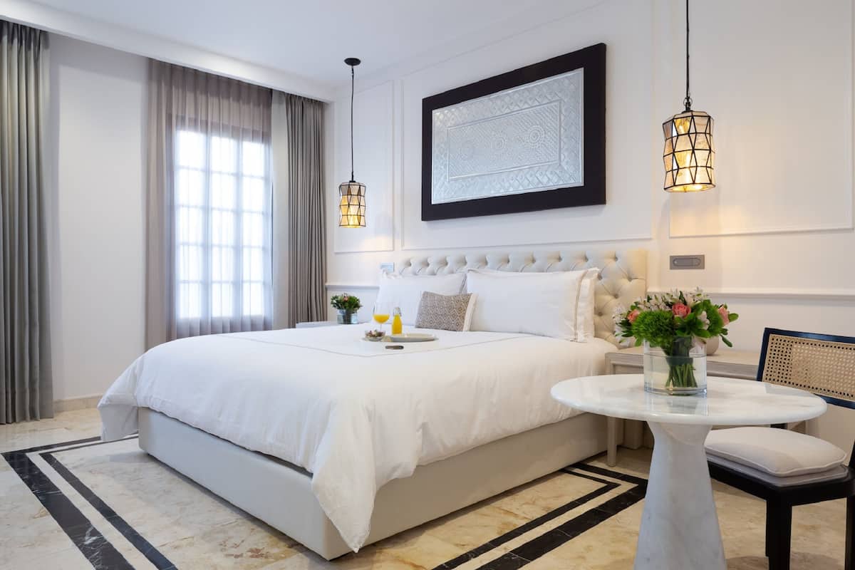 Una habitación de hotel moderna con una cama grande, una mesa redonda y una silla. Se aprecia una obra de arte enmarcada en la pared y una ventana con cortinas.