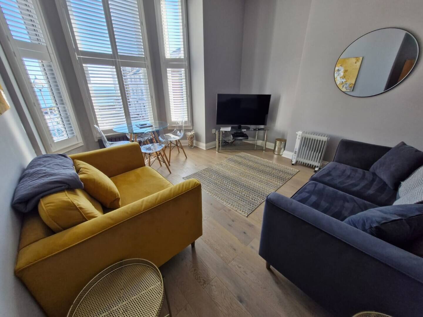 A modern living room with a yellow sofa, a dark sofa, a TV, and a dining area with a glass table and chairs.
