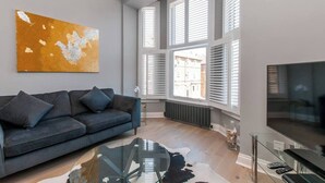 A modern living room with a grey sofa, a glass coffee table, and a large window with blinds. - Serenity by the Sea - Broadstairs (Kent)