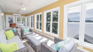 A sunlit room with wicker furniture, large windows, and a view of a body of water. - MAIN LAKE VIEWS - Fire Pit - Kayaks, SUPs - Pool Table - Bikes (Littleton)