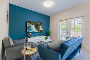 A modern living room with a blue accent wall, a large flat-screen TV, a grey sofa, a blue armchair, and a coffee table with a bottle and snacks.