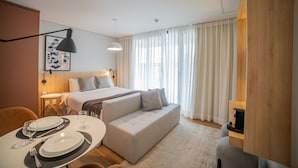 A modern hotel room with a bed, sofa, dining table set with plates and glasses, and a large window with curtains.
