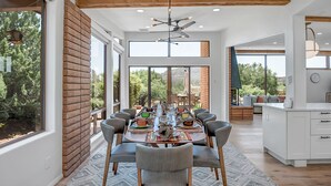A spacious dining area with a brick accent wall, large windows, and a ceiling fan. - Resort Style Retreat, Red Rock Views Inside & Out+Salt Water Pool+Hot Tub (Sedona)