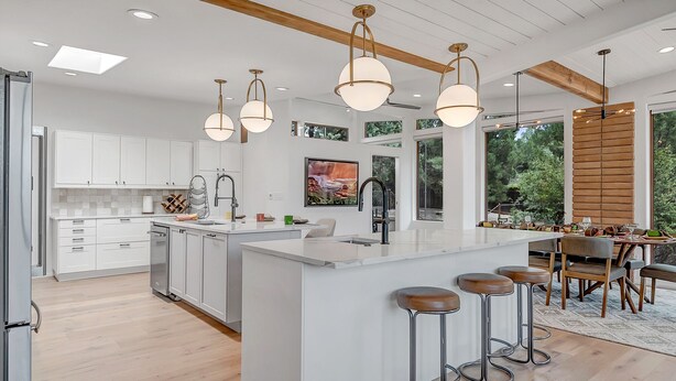 A modern kitchen with white cabinetry, a central island with stools, pendant lighting, and a dining area with a view of greenery. - Resort Style Retreat, Red Rock Views Inside & Out+Salt Water Pool+Hot Tub (Sedona)
