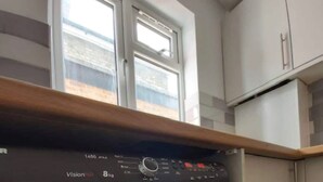 A front-loading washing machine with a control panel, located in a kitchen with a window above it. - Orpington RSG House (Orpington)