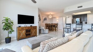 A modern living room with a large white sofa, a dining area with a wooden table and chairs, a kitchen with dark cabinets, and a flat-screen TV mounted on the wall.