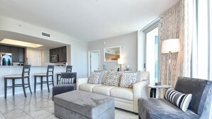 A modern living room with a grey ottoman, a sofa with patterned cushions, and a dark armchair. A kitchen with dark cabinets and a microwave is visible in the background.