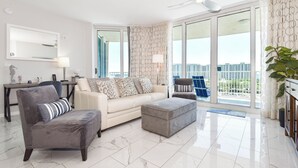 A modern living room with a sofa, armchair, and ottoman, a dining area with a table and chairs, and large windows with a city view.