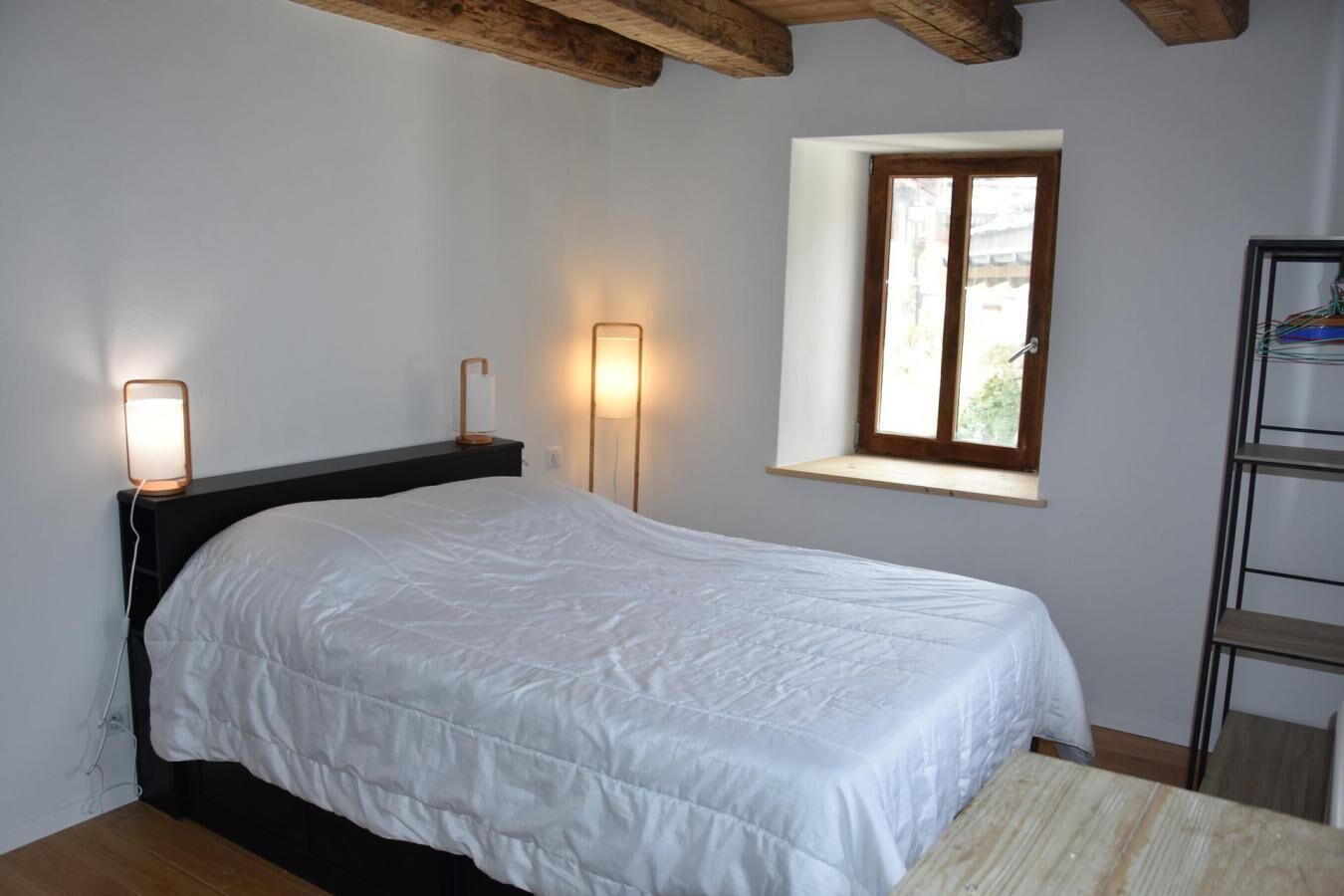A bedroom with a bed, bedside lamps, a window, and a wooden beam ceiling.