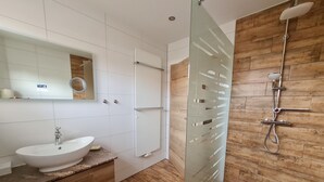 Un baño moderno con piso de madera, un lavamanos blanco sobre encimera de mármol, cabina de ducha con paredes de vidrio y un panel de pared de madera.