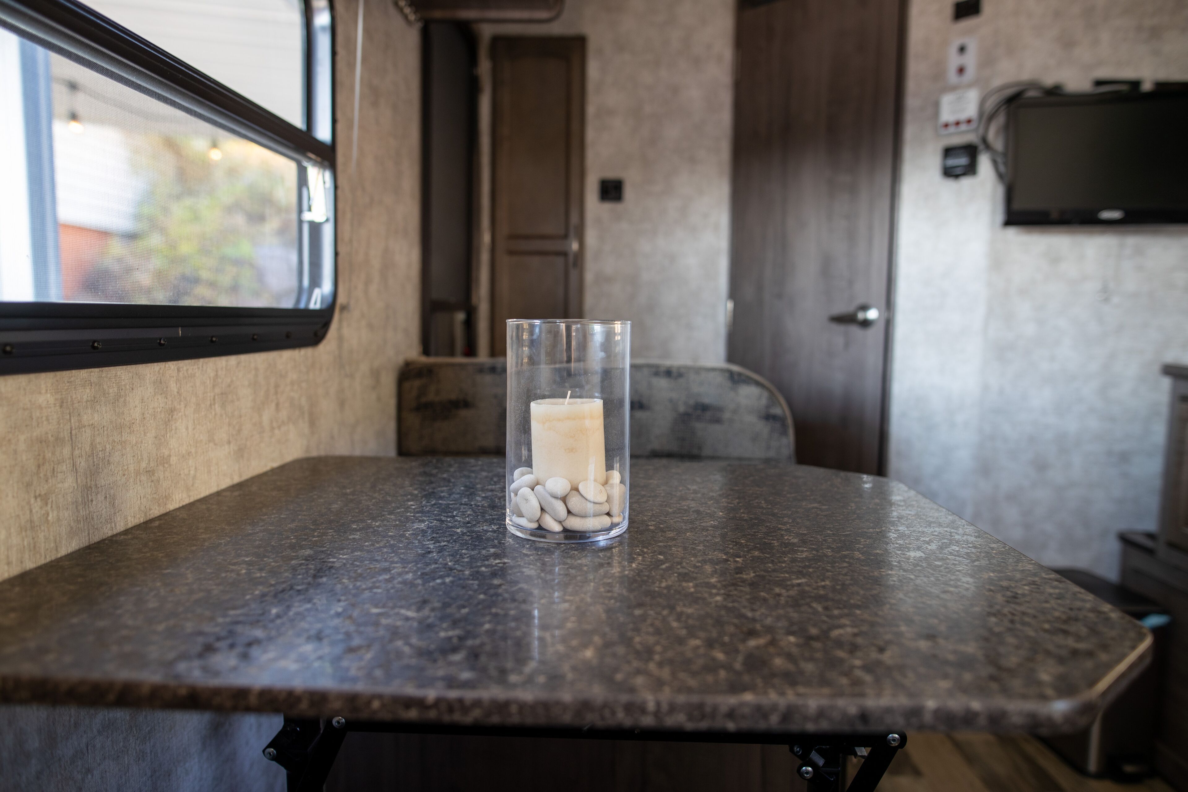 A table with a glass containing stones and a candle, a window, and a door in a room with a textured wall.