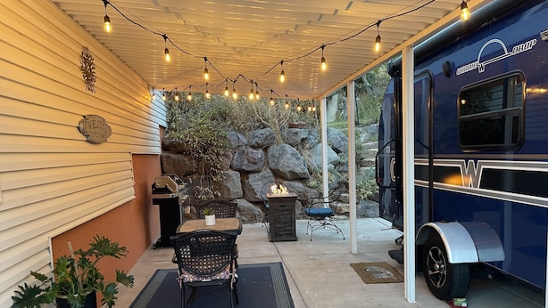A covered patio with string lights, a grill, a fire pit, and a blue RV. - Charming Winnie Drop Trailer with Cozy Private Patio Escape (Farmington)