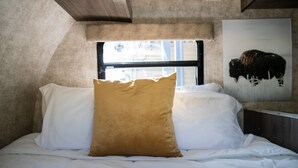 A bed with white and beige pillows, a window with a view of buildings, and a wall-mounted painting of a bison. - Charming Winnie Drop Trailer with Cozy Private Patio Escape (Farmington)