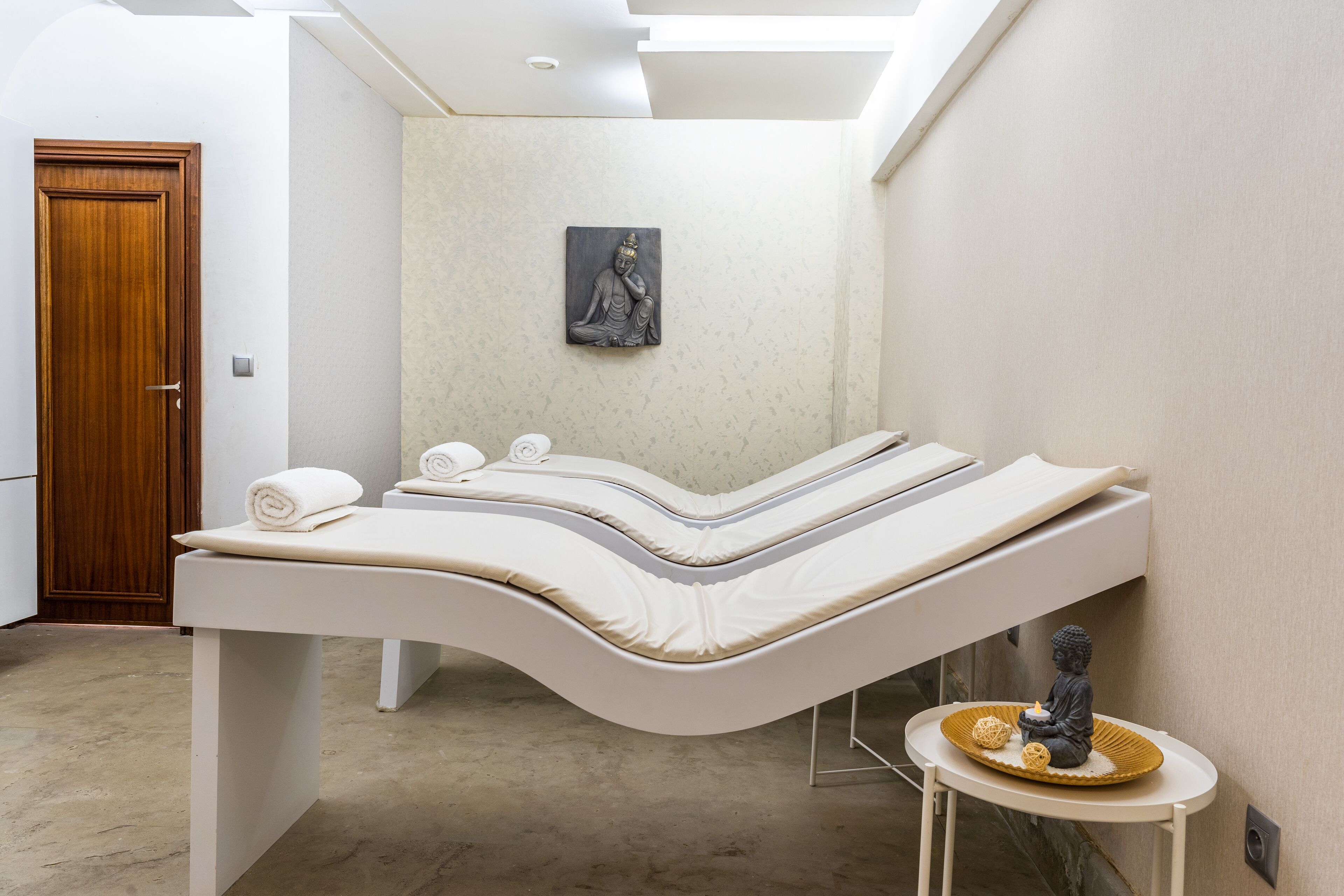 A modern spa room with a curved massage table, a small statue, and a wall-mounted artwork.