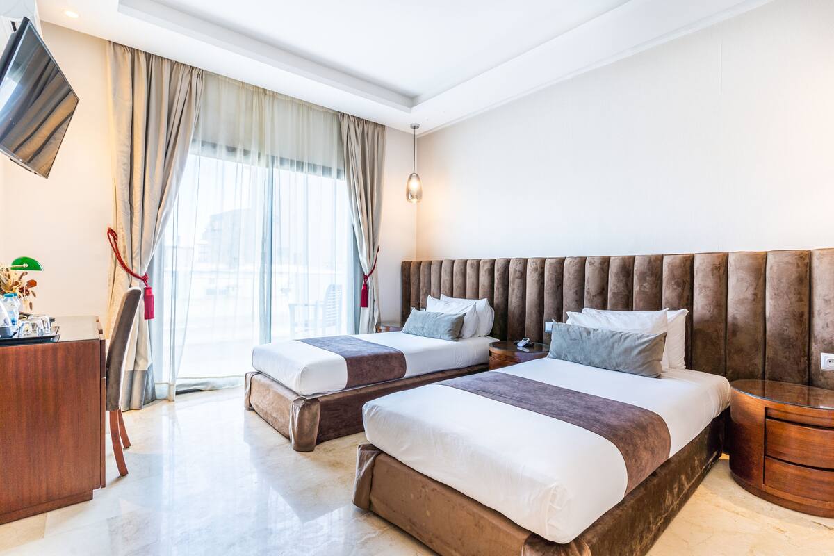 Habitación de hotel con dos camas, un televisor montado en la pared, un escritorio y un ventanal con cortinas.