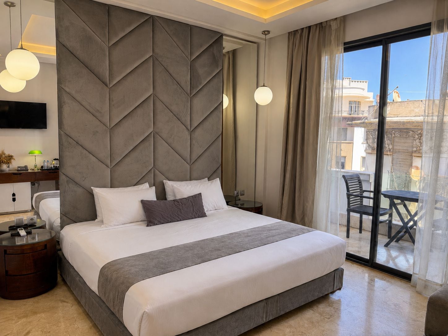 Ein modernes Hotelzimmer mit einem großen Bett, einem Flachbildfernseher, einem Nachttisch mit Lampe und Blick auf die Stadt durch ein großes Fenster.