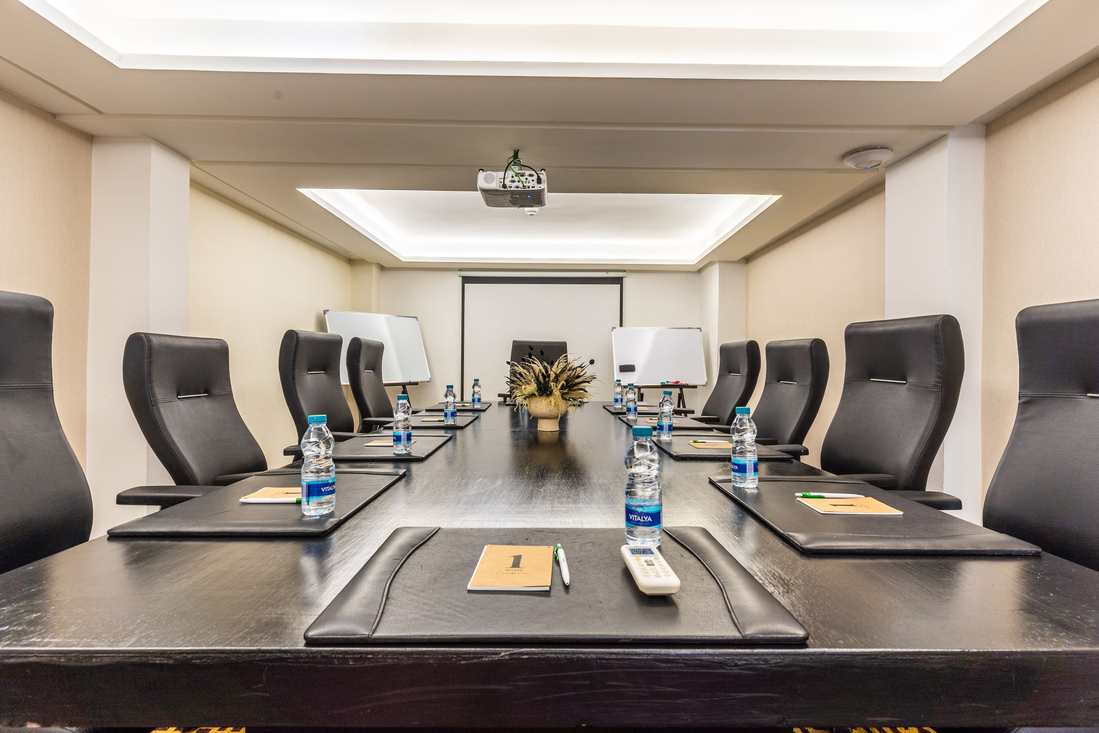 Une salle de conférence avec une longue table, des chaises noires, des bouteilles d’eau, un projecteur et une petite plante.