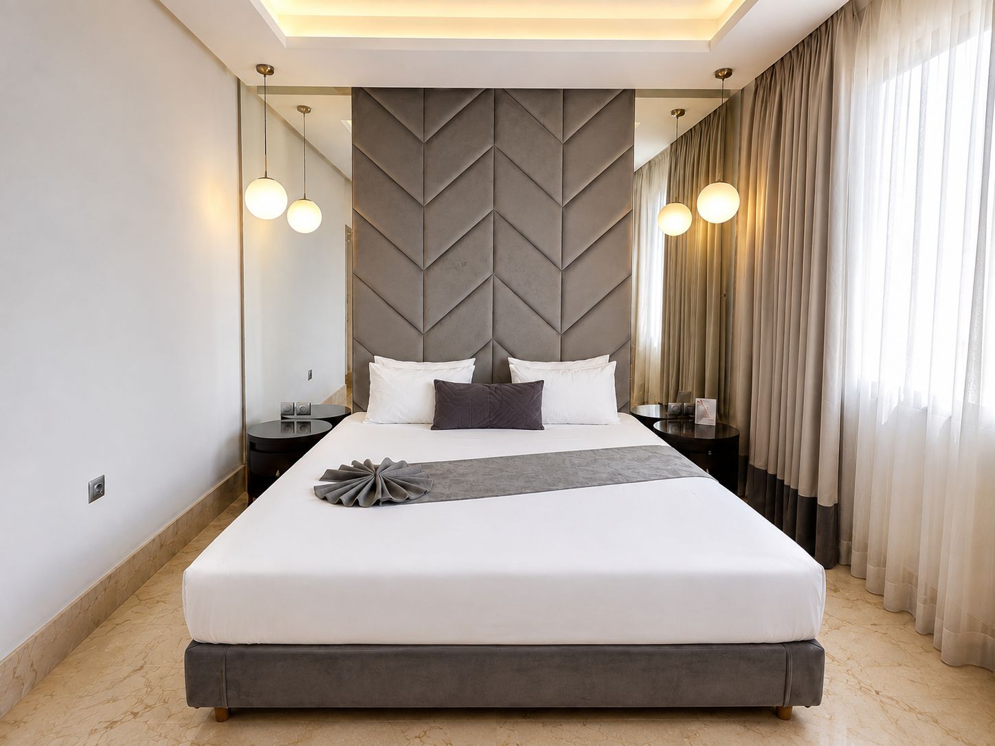 Ein modernes Hotelzimmer mit einem großen Bett, einem dekorativen Kopfteil, Nachttischen und Blick auf die Stadt durch transparente Vorhänge.