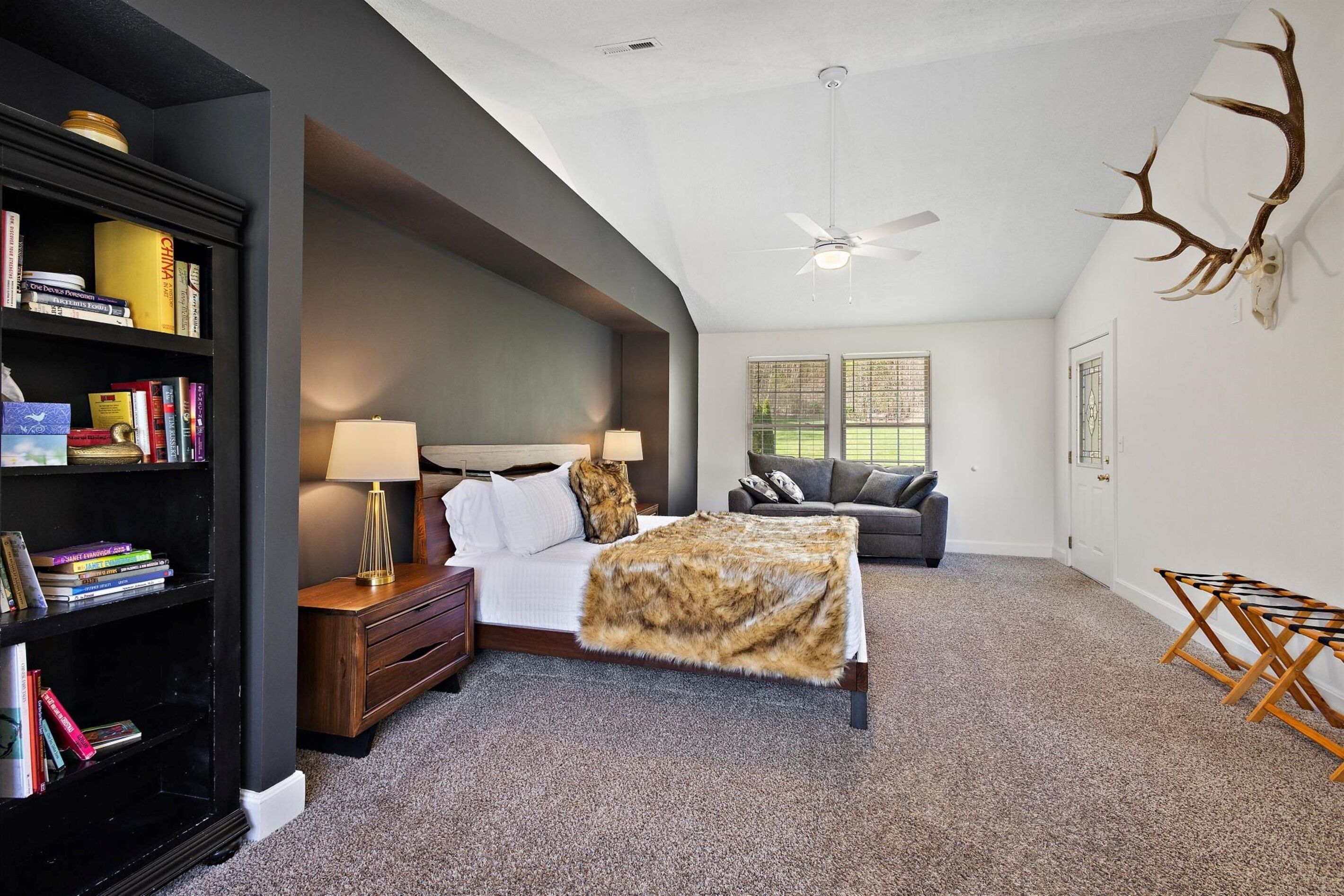 A bedroom with a bed, a bookshelf, a desk, a lamp, a grey sofa, and a deer antler decoration.