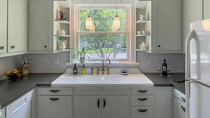 Une cuisine avec des meubles blancs, un plan de travail blanc, une fenêtre donnant sur un espace verdoyant et un réfrigérateur blanc.