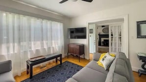 Un salon avec un canapé gris, un banc, un tapis à motifs et un ventilateur de plafond.