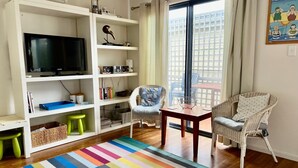 Een woonkamer met een televisie, een boekenkast, rieten stoelen, een kleurrijk gestreept tapijt en een uitzicht door een groot raam.