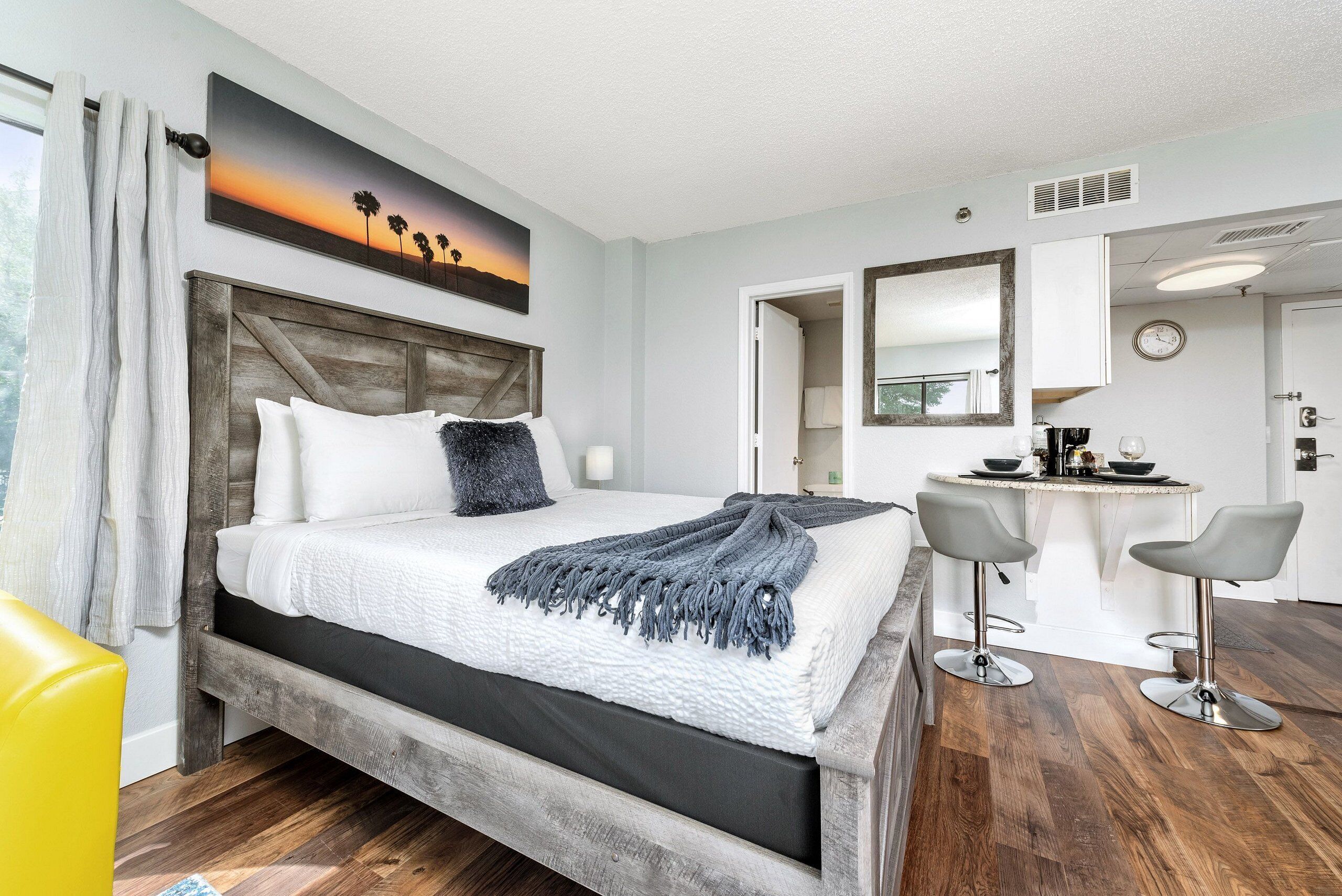 Quarto de hotel com cama, escrivaninha e duas cadeiras, um quadro na parede e um relógio.