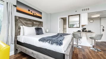 Quarto de hotel com cama, escrivaninha e duas cadeiras, um quadro na parede e um relógio.